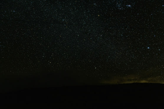 A dark night sky filled with countless stars above a barely visible landscape; a faint line, possibly a power line, stretches horizontally across the top part of the image. by Justin Salem Meyer
