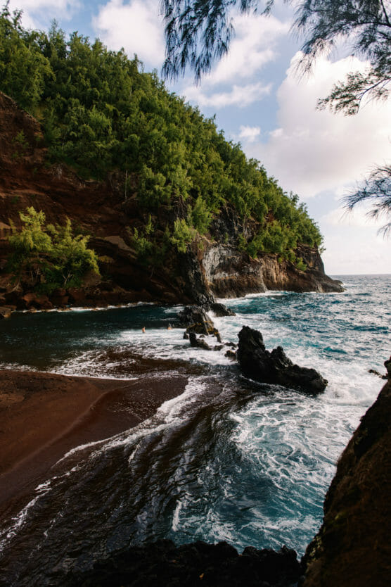 A rugged coastline with lush green trees atop red cliffs, waves crashing against black rocks, and a narrow strip of dark sandy beach under a partly cloudy sky. by Justin Salem Meyer