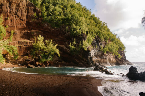 A secluded beach with dark, reddish-brown sand, rocky outcrops, and lush green trees on steep cliffs, with waves crashing along the shoreline under a partly cloudy sky. by Justin Salem Meyer