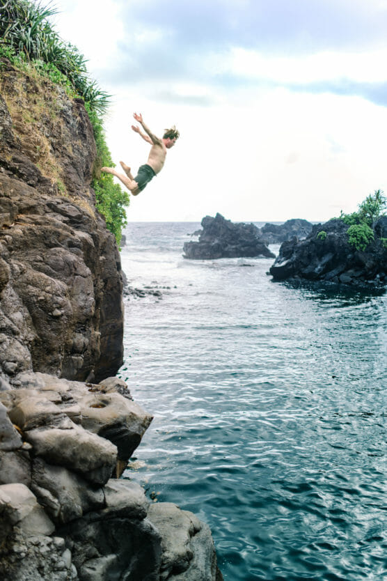 A person wearing swim trunks leaps off a rocky cliff into the blue ocean below, surrounded by jagged rocks and lush greenery, under a cloudy sky. by Justin Salem Meyer