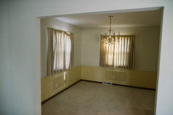 An empty room with cream-colored walls, beige carpet, two curtained windows, a central chandelier, and decorative molding on the lower walls. Sunlight filters softly through the curtains. by Justin Salem Meyer