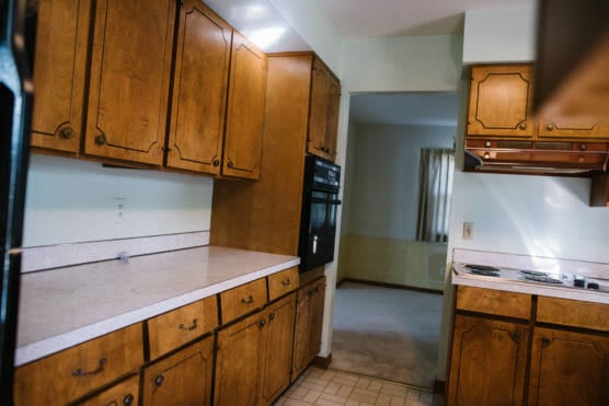 A kitchen with wooden cabinets, white countertops, a built-in oven, and an electric stovetop. The floor has a tiled pattern, and an adjacent room with carpet and a window with beige curtains is visible. by Justin Salem Meyer
