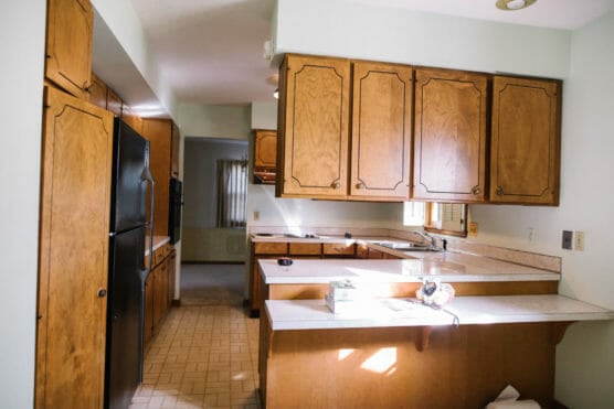 A kitchen with wooden cabinets, white countertops, a black refrigerator, and a stove. Sunlight streams through a window above the sink, and some items are on the counters. The floor has a light-colored tile pattern. by Justin Salem Meyer