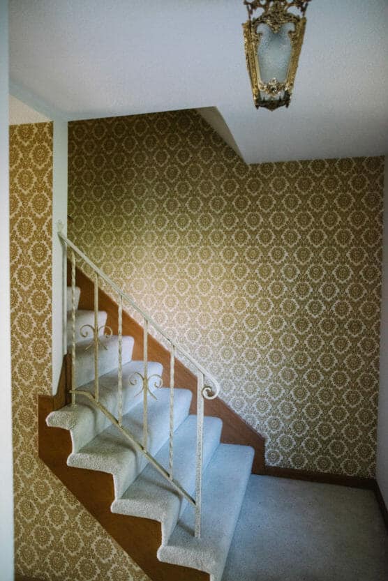 A staircase with white carpeting and an ornate metal railing, set against a wall covered in vintage, gold-patterned wallpaper. An antique light fixture hangs from the white ceiling above. by Justin Salem Meyer