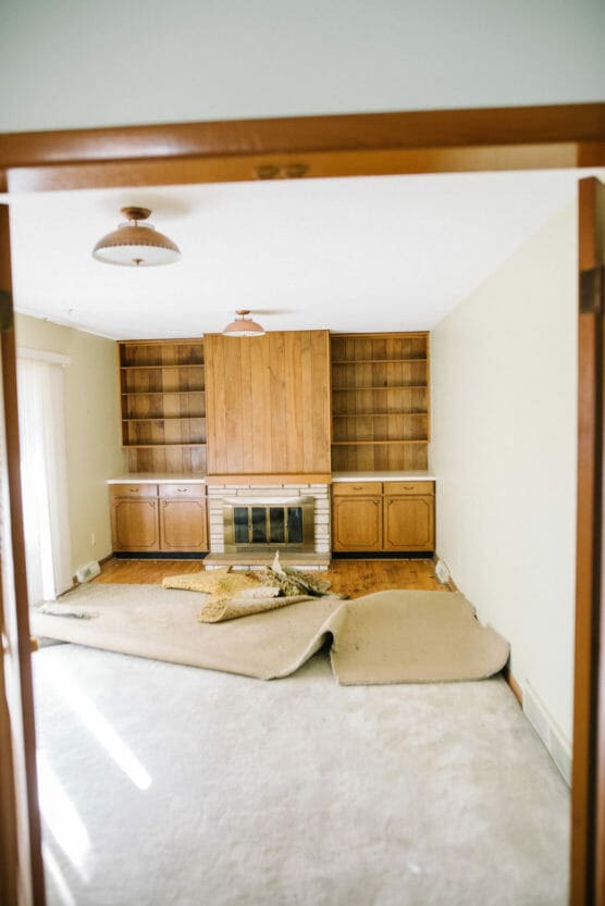 A living room with wood built-in shelves and a fireplace, old carpet partially pulled up, exposing the floor; light streams in from the left, and uncushioned light fixtures hang from the ceiling. by Justin Salem Meyer