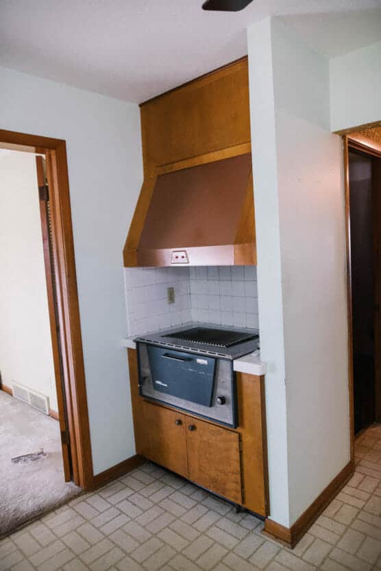 A vintage built-in oven with a copper hood in a wood-paneled kitchen nook, set against white tiled walls and light-colored flooring. An open doorway and carpeted room are visible to the left. by Justin Salem Meyer