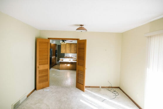 A bright, empty room with beige carpet and walls, open wooden double doors leading to a kitchen with wooden cabinets. Sunlight enters through vertical blinds on the right; some cables are on the floor. by Justin Salem Meyer