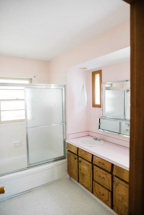 A bathroom with light pink walls, a wooden vanity with pink countertop, built-in sink, mirrored cabinet, pendant light, frosted glass shower door, and a window above the bathtub. by Justin Salem Meyer