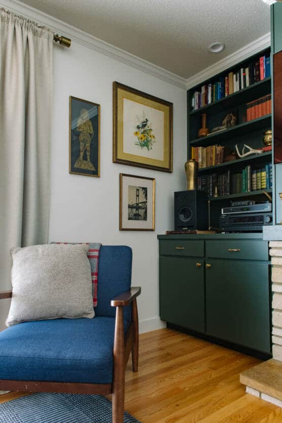 A cozy living room corner with a blue armchair and gray pillow, green cabinets with gold knobs, shelves filled with books and decor, framed artwork on a white wall, and light wood flooring. by Justin Salem Meyer