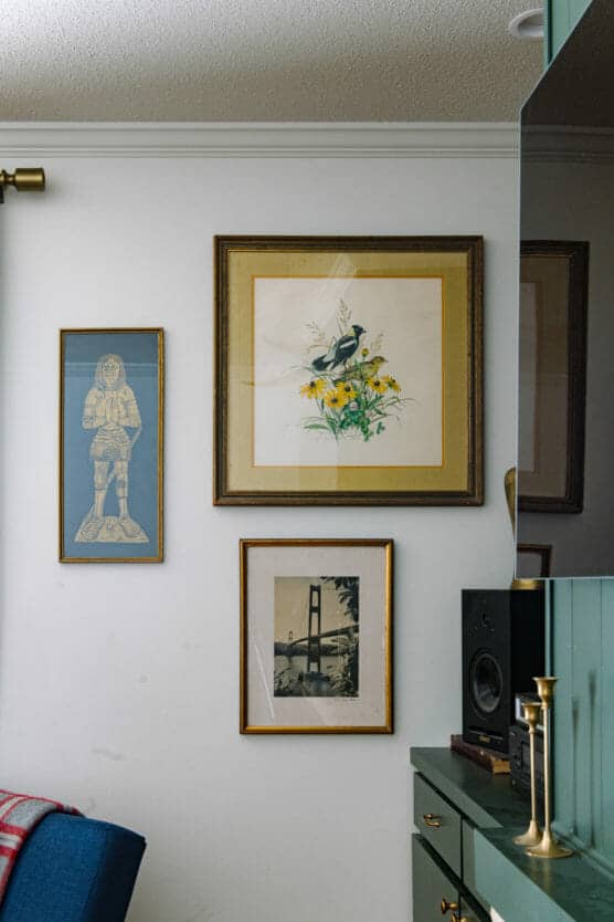 A wall displays three framed artworks: a blue-toned figure, a floral painting with two birds, and a black-and-white photo of a bridge, above a green cabinet with a speaker, brass candlestick, and small decor items. by Justin Salem Meyer