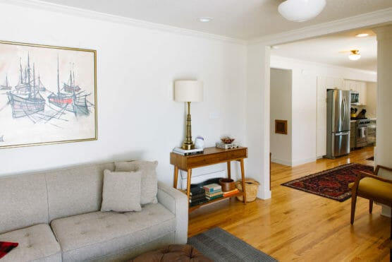 A bright living room with a light gray sofa, a framed ship painting on the wall, a wooden console table with a lamp, and a view into the kitchen with stainless steel appliances and hardwood floors. by Justin Salem Meyer