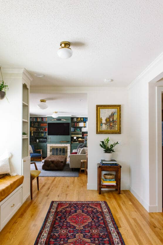 A cozy living space with hardwood floors, a colorful rug, bookshelves, a fireplace, and a seating area. A framed painting and a side table with plants and books are visible in the bright, inviting room. by Justin Salem Meyer