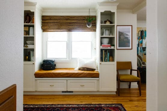 A cozy reading nook with a cushioned bench under a window, flanked by built-in shelves with books and decor. There’s a brown chair, framed art, and a bag on the bench. Hardwood floors and a patterned rug complete the space. by Justin Salem Meyer