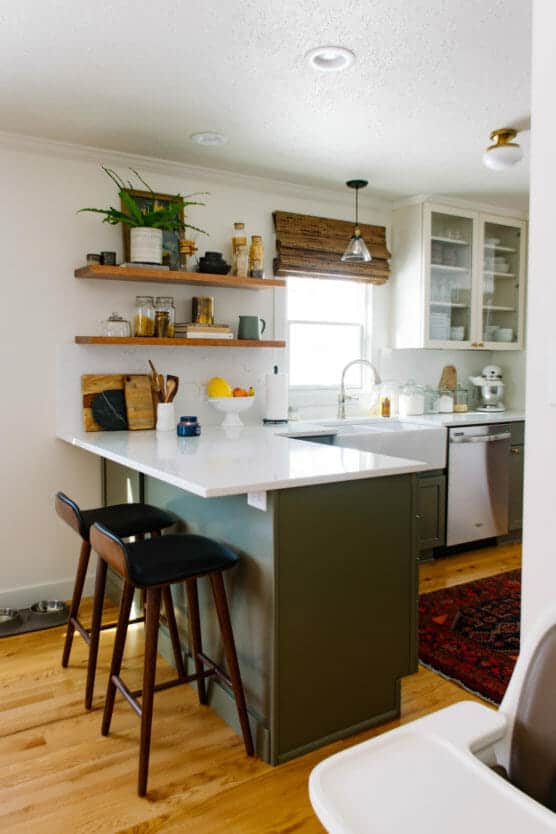 A modern kitchen with green lower cabinets, white countertops, open wooden shelves with dishes and plants, two barstools, a window with a bamboo shade, and stainless steel appliances on hardwood floors. by Justin Salem Meyer