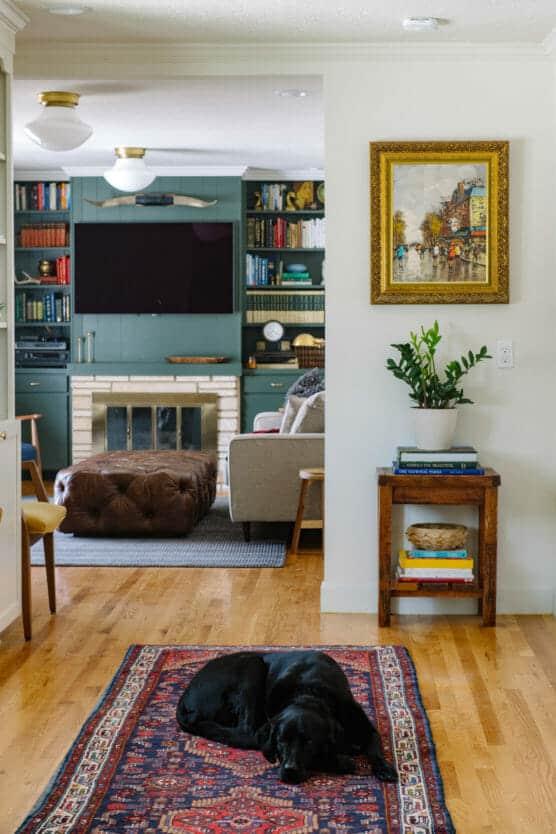 A black dog sleeps on a patterned rug in a hallway with hardwood floors. In the background, a cozy living room features a fireplace, built-in bookshelves, a TV, and a painting on the wall above a small table with a plant. by Justin Salem Meyer