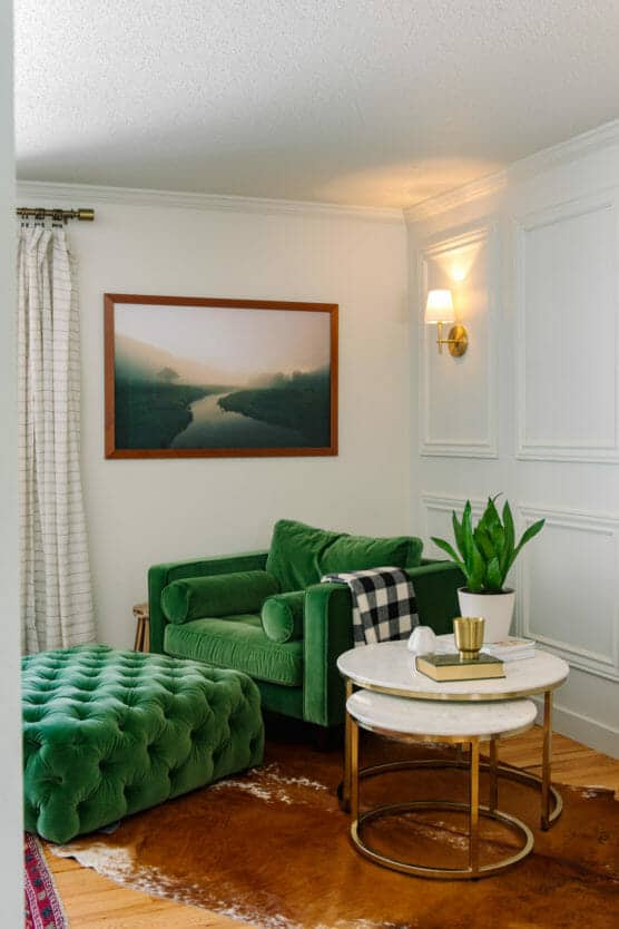 A cozy corner with a green velvet armchair and matching ottoman, a round marble coffee table with a plant and books, a wall sconce, a landscape photo, white paneled walls, and a patterned rug on wood floors. by Justin Salem Meyer