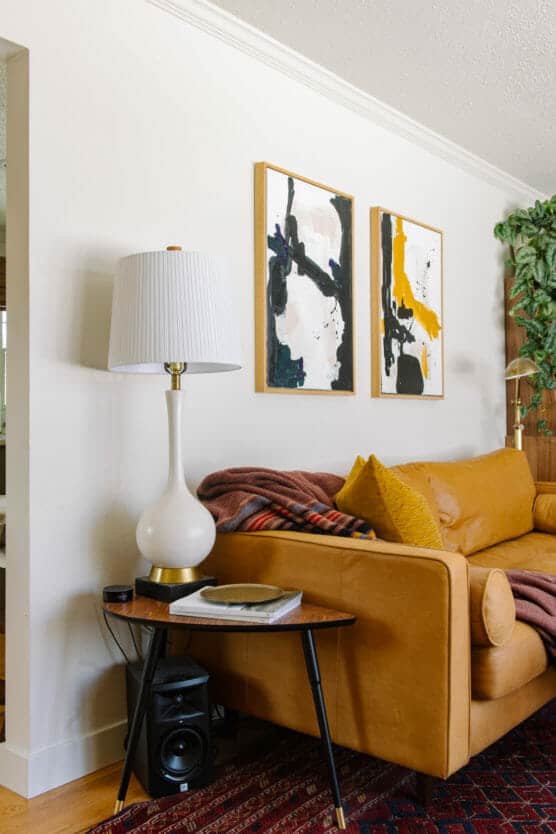 A cozy living room with a tan leather sofa, mustard cushion, and a colorful blanket. A side table holds a lamp and record player. Abstract art hangs on the white wall above the sofa, and a green plant is in the corner. by Justin Salem Meyer