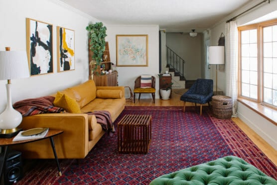A cozy living room with a tan sofa, abstract art, green ottoman, dark blue chair, red patterned rug, wooden furniture, large window with curtains, houseplants, and light fixtures. Stairs are visible in the background. by Justin Salem Meyer