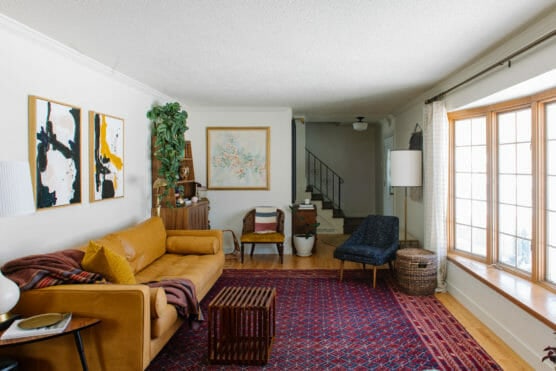 A cozy living room with a tan leather sofa, dark blue chair, red patterned rug, large window with sheer curtains, abstract wall art, houseplants, and a wooden sideboard near a staircase. by Justin Salem Meyer
