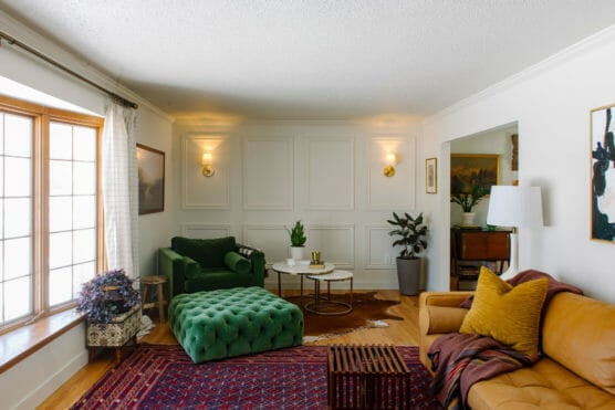 A cozy living room with a green armchair and ottoman, a tan leather sofa with yellow cushions, a large window, houseplants, two round coffee tables, and patterned rugs on a wooden floor. Warm lighting and wall art decorate the space. by Justin Salem Meyer