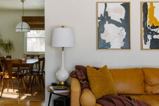 A cozy living room with a yellow sofa, gold pillow, and striped blanket, next to a white lamp and side table. Abstract art hangs on the wall. A dining area with wooden chairs and a hanging light is seen in the background. by Justin Salem Meyer