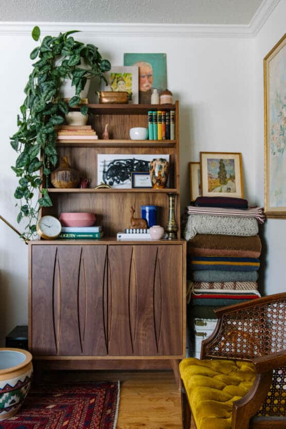 A wooden cabinet with shelves displaying books, plants, vases, and decor sits next to a tall stack of folded blankets. A yellow velvet chair and a patterned rug are partially visible in the cozy, eclectic room. by Justin Salem Meyer
