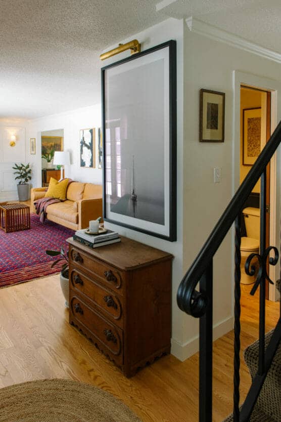 A cozy living room with a yellow sofa, colorful rug, wall art, wooden side table with books and a mug, and a staircase with black railing leading to another room. Natural light fills the space. by Justin Salem Meyer