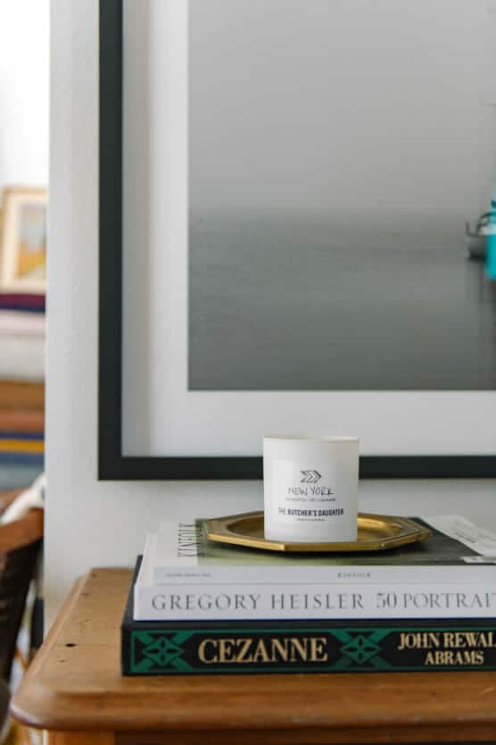 A white candle labeled THE BUTCHERS DAUGHTER sits on a brass tray atop a stack of books, including 50 Portraits by Gregory Heisler and Cezanne. A framed photograph of a boat on water hangs behind. by Justin Salem Meyer