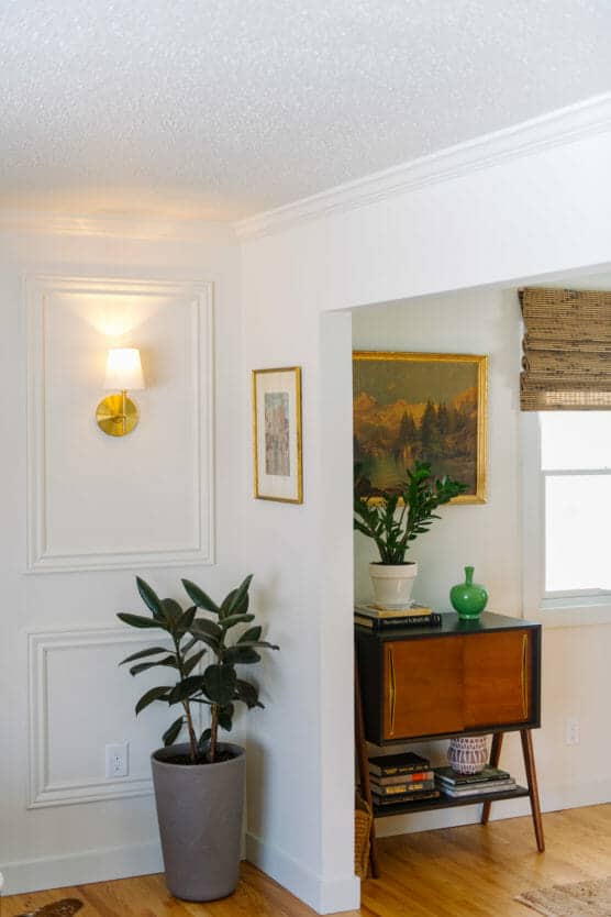 A bright living room corner featuring a potted plant, wall-mounted sconce, framed artwork, wooden side table with books, green vase, potted plant, and a landscape painting above it. Hardwood floors and white walls. by Justin Salem Meyer