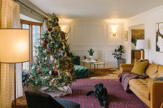 A cozy living room with a decorated Christmas tree, a yellow sofa, green ottoman, red rug, and a black dog lying on the floor. Warm lighting and festive decor create a welcoming holiday atmosphere. by Justin Salem Meyer