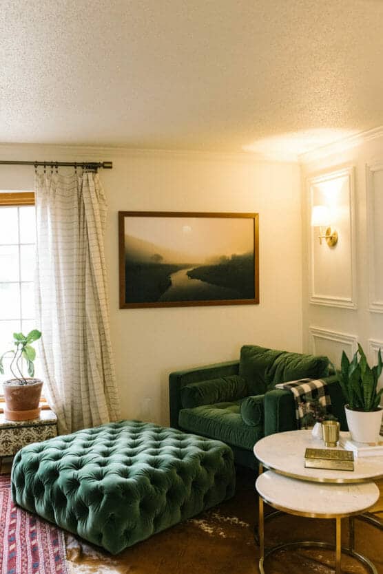 Cozy living room with a green velvet armchair, matching tufted ottoman, marble side tables, potted plants, striped curtains, and a landscape photo on the wall above the chair. Warm lighting creates a welcoming atmosphere. by Justin Salem Meyer