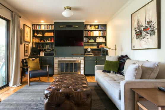 A cozy living room with a beige sofa, a tufted leather ottoman, an armchair, a fireplace under a wall-mounted TV, built-in bookshelves, and nautical artwork on the wall. Warm lighting and wood floors complete the space. by Justin Salem Meyer
