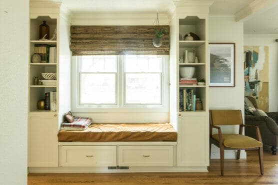 A cozy window seat with a mustard cushion and plaid blanket is built between white shelves filled with books and decor. A chair sits nearby, and sunlight streams through a window with a bamboo shade. by Justin Salem Meyer