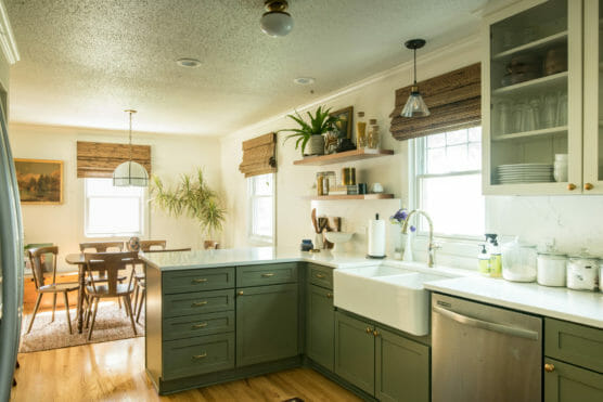 A bright kitchen with sage green cabinets, a white farmhouse sink, marble countertops, open shelves with plants and dishes, and a dining area with a wooden table in the background. Natural light fills the space. by Justin Salem Meyer