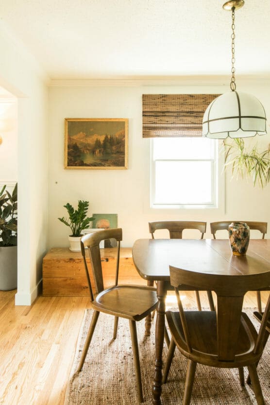 Bright dining room with a wooden table and four chairs on a woven rug, a pendant light above, a potted plant on a wooden chest, wall art, and a window with a bamboo shade letting in natural light. by Justin Salem Meyer