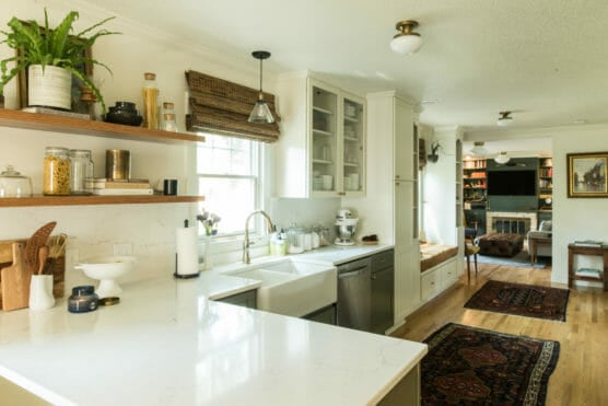 Bright kitchen with white countertops, farmhouse sink, open shelves with jars and plants, window with bamboo shade, and view into a cozy living room with rugs, bookshelves, and a fireplace. by Justin Salem Meyer