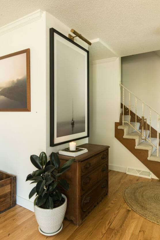 A wooden chest with a potted plant, a candle, and books sits against a white wall. Above it hangs a framed photo of a sailboat, lit by a brass picture light. A staircase and woven rug are nearby. by Justin Salem Meyer