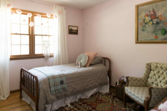 A cozy bedroom with a single bed, pastel bedding, and decorative pillows sits by a window with sheer curtains. The room features a patterned rug, an armchair, soft wall colors, and floral artwork above the bed. by Justin Salem Meyer