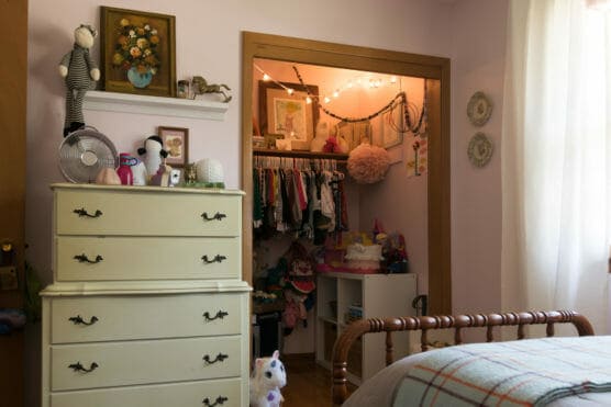 A cozy, softly lit childs bedroom with a cream dresser, stuffed animals, and a closet filled with clothes and toys. String lights decorate the closet, and a plush unicorn sits on the floor. by Justin Salem Meyer