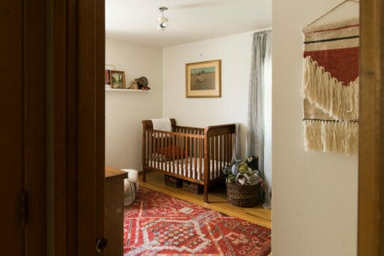 A cozy nursery with a wooden crib, stuffed animals, a red patterned rug, a shelf with books and decor, framed art on the wall, and a woven wall hanging near the door. Soft light enters through a curtained window. by Justin Salem Meyer