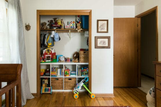 A neatly organized childs closet with clothes hanging and shelves holding toys, books, and wicker baskets. A small walker toy is in front of the closet. Sunlight enters from a window on the left, brightening the wooden floor. by Justin Salem Meyer