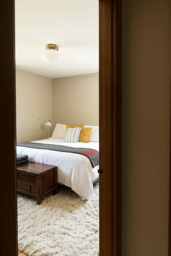 A cozy bedroom viewed through a doorway, featuring a neatly made bed with white bedding, yellow and striped pillows, a wooden chest at the foot, a fluffy rug, and a wall-mounted lamp. by Justin Salem Meyer