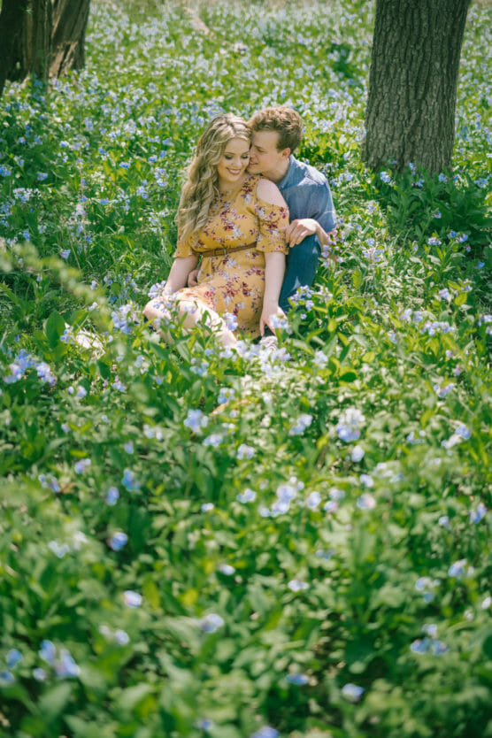 A couple sits closely together in a field of green grass and purple wildflowers. The woman wears a yellow floral dress, and the man sits behind her, leaning in affectionately. Sunlight filters through the trees. by Justin Salem Meyer