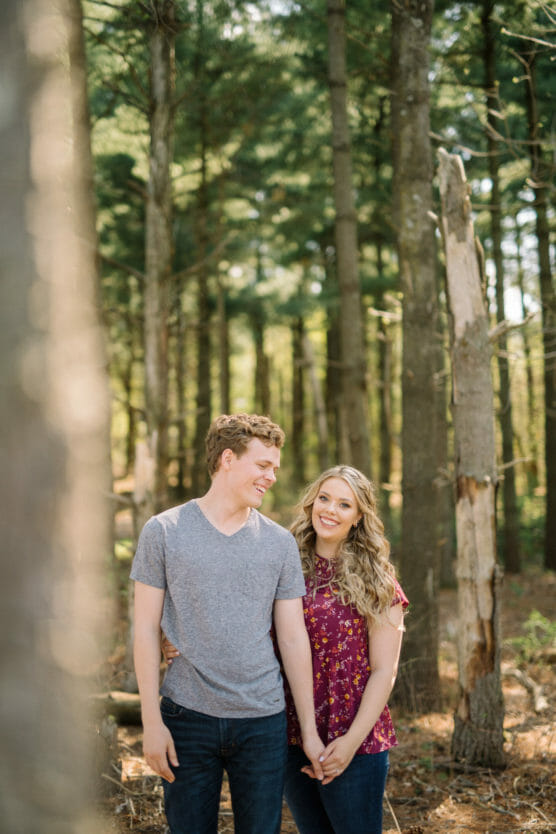 A young couple stands in a sunlit forest, holding hands and smiling at each other. The man wears a gray t-shirt and jeans, while the woman wears a purple floral top and jeans. Tall trees surround them in the background. by Justin Salem Meyer