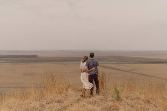 A couple stands on a grassy hill, embracing as they look out over a vast, dry landscape under an overcast sky. The scene feels peaceful and expansive. by Justin Salem Meyer