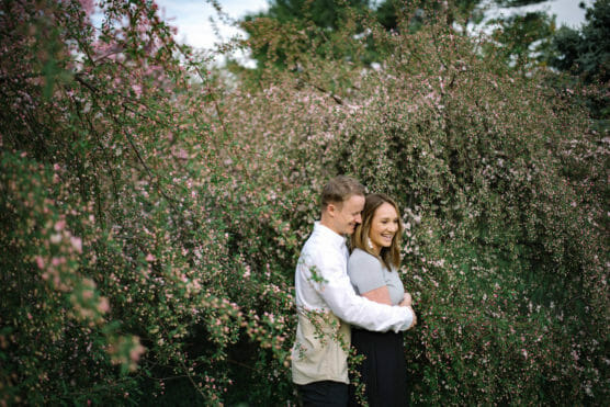 A couple stands closely together, smiling and embracing in front of dense, blossoming bushes with small pink flowers on a cloudy day. by Justin Salem Meyer