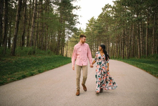 A couple walks hand in hand down a paved road surrounded by tall trees. The man wears a pink shirt and beige pants; the woman wears a long floral dress. They look at each other and smile. by Justin Salem Meyer