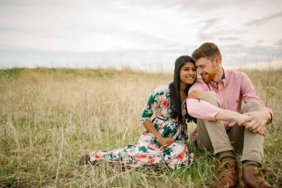 A couple sits closely together in a grassy field. The woman wears a floral dress and leans on the mans shoulder, smiling. The man, in a pink shirt and khaki pants, smiles warmly. The sky is lightly clouded. by Justin Salem Meyer