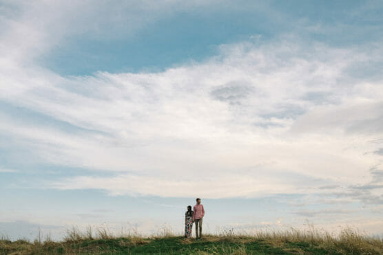 Two people stand together on a grassy hill under a vast, partly cloudy sky, with the horizon low in the frame and wide open space above them. by Justin Salem Meyer