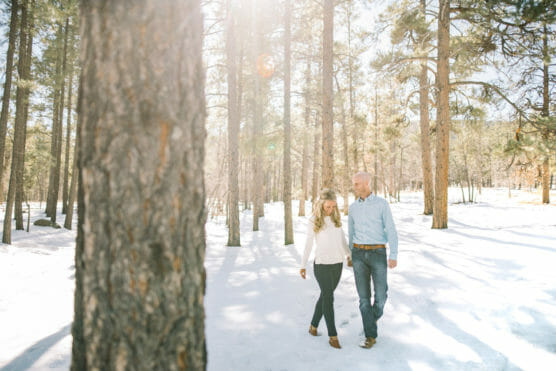 A couple walks hand in hand through a snowy forest with sunlight streaming through tall pine trees. Both are casually dressed and appear happy, surrounded by bright, wintry scenery. by Justin Salem Meyer